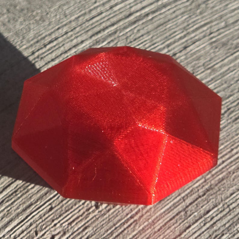 Op de afbeelding: Een facetrijk, rood, geometrisch object met een glanzend oppervlak. Het object heeft een veelzijdige vorm, met vlakke vlakken die elkaar in scherpe hoeken ontmoeten. De kleur is een levendig, effen rood en het oppervlak reflecteert licht.