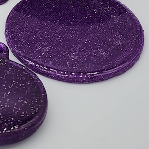 K&ouml;nnte beinhalten: Zwei runde, violette, glitzernde Harzst&uuml;cke. Das gr&ouml;&szlig;ere St&uuml;ck ist leicht erh&ouml;ht und hat eine glatte, gl&auml;nzende Oberfl&auml;che. Das kleinere St&uuml;ck ist flach und hat eine texturreichere Oberfl&auml;che.