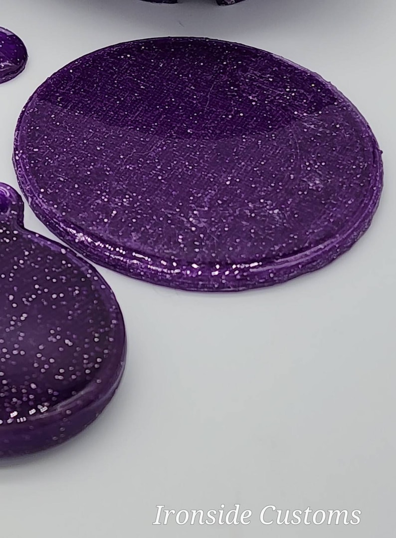 K&ouml;nnte beinhalten: Zwei runde, violette, glitzernde Harzst&uuml;cke. Das gr&ouml;&szlig;ere St&uuml;ck ist leicht erh&ouml;ht und hat eine glatte, gl&auml;nzende Oberfl&auml;che. Das kleinere St&uuml;ck ist flach und hat eine texturreichere Oberfl&auml;che.