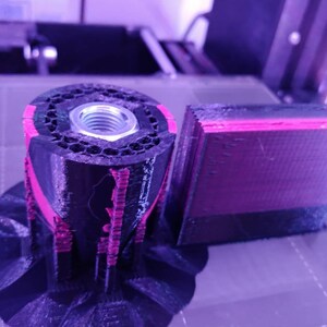 Op de afbeelding: Een 3D-geprint object in zwart en roze, met een zeshoekig patroon en een metalen moer. Het object bestaat uit meerdere lagen, met een apart rechthoekig stuk ernaast. De afbeelding is gemaakt in een werkplaats.