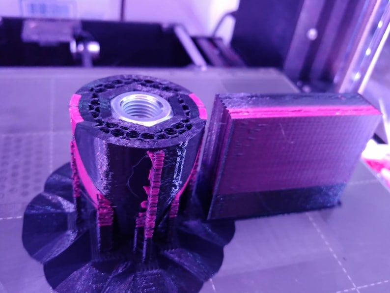 Op de afbeelding: Een 3D-geprint object in zwart en roze, met een zeshoekig patroon en een metalen moer. Het object bestaat uit meerdere lagen, met een apart rechthoekig stuk ernaast. De afbeelding is gemaakt in een werkplaats.