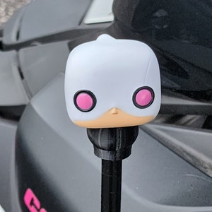 May include: White figure with pink eyes and a black base, resembling a character from a popular cartoon. The figure is attached to a black stick, likely a car antenna topper.
