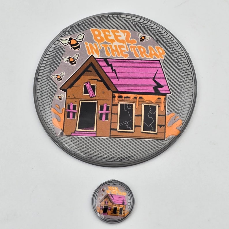 K&ouml;nnte beinhalten: Ein runder grauer Pin mit einem Cartoon-Haus mit einem rosafarbenen Dach und tropfendem gelbem Anstrich. Das Haus hat zerbrochene Fenster und eine T&uuml;r. Der Text "Beez in the Trap" befindet sich &uuml;ber dem Haus. Um das Haus herum befinden sich Cartoon-Bienen.