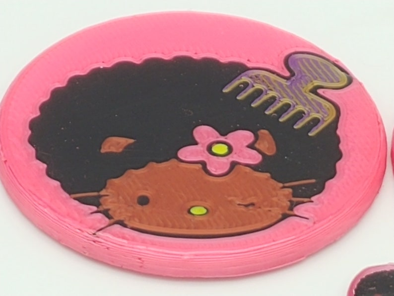 K&ouml;nnte beinhalten: Runder rosa Untersetzer mit einem schwarzen und braunen Hello Kitty-Charakter mit Afro-Frisur, einer rosa Blume und einem lila und goldenen Kamm.
