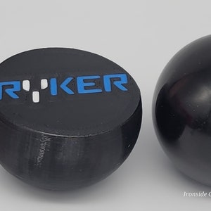 Op de afbeelding: Twee zwarte, bolvormige objecten. E&eacute;n is een afgeplatte bol met het woord "RYKER" in blauwe en witte letters. De andere is een effen zwarte bol. De objecten staan tegen een lichtgrijze achtergrond.