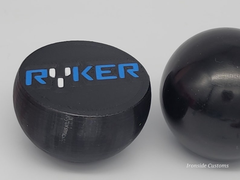 Op de afbeelding: Twee zwarte, bolvormige objecten. E&eacute;n is een afgeplatte bol met het woord "RYKER" in blauwe en witte letters. De andere is een effen zwarte bol. De objecten staan tegen een lichtgrijze achtergrond.