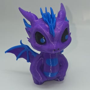 Op de afbeelding: Een paars en blauw drakenbeeldje. De draak heeft grote zwarte ogen met blauwe pupillen, blauwe accenten op zijn hoofd, vleugels en lichaam. Het beeldje is een grillig, fantasie-thema decoratief item.
