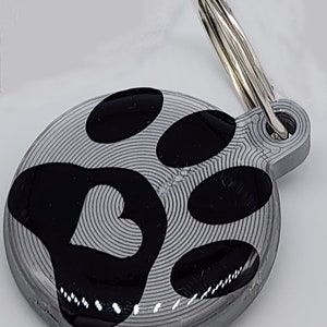 May include: A silver dog tag with a black paw print design. The paw print has a heart shape in the center.