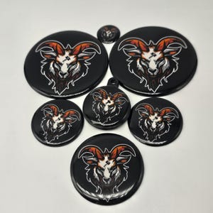 May include: A collection of seven circular black acrylic or plastic discs of varying sizes, each featuring a printed graphic of a goat head with orange and white horns, a white face, and dark fur, with one disc including a small hanging loop.