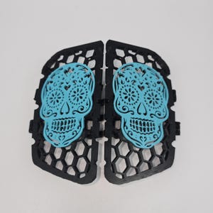 May include: Two decorative panels featuring a Day of the Dead sugar skull design. The skulls are a vibrant turquoise color and are set against a black, honeycomb-patterned background. The panels are symmetrical and designed to be displayed together.