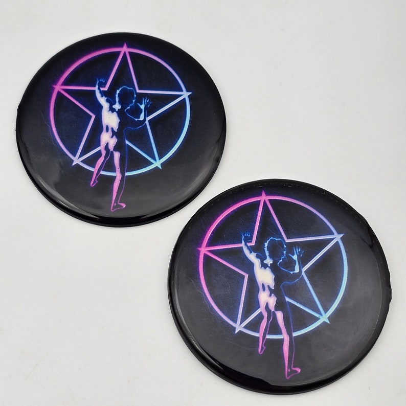 K&ouml;nnte beinhalten: Zwei schwarze runde Kn&ouml;pfe mit einem pinkfarbenen und blauen Pentagramm-Design. Eine Silhouette einer Person befindet sich im Pentagramm.