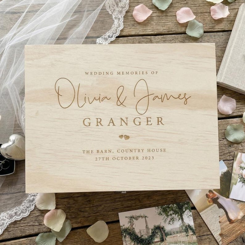 May include: A rectangular light-colored wooden keepsake box featuring the engraved text WEDDING MEMORIES OF Olivia & James GRANGER, two small heart icons, THE BARN, COUNTRY HOUSE, and the date 27TH OCTOBER 2023 centered on the lid.