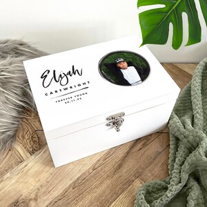 Könnte beinhalten: Eine weiße Holz-Andenkenbox mit silbernem Verschluss. Der Deckel zeigt ein rundes Foto einer Person, den Namen "Elijah" in Schreibschrift und den Text "CARTWRIGHT FOREVER YOUNG 20.11.25".