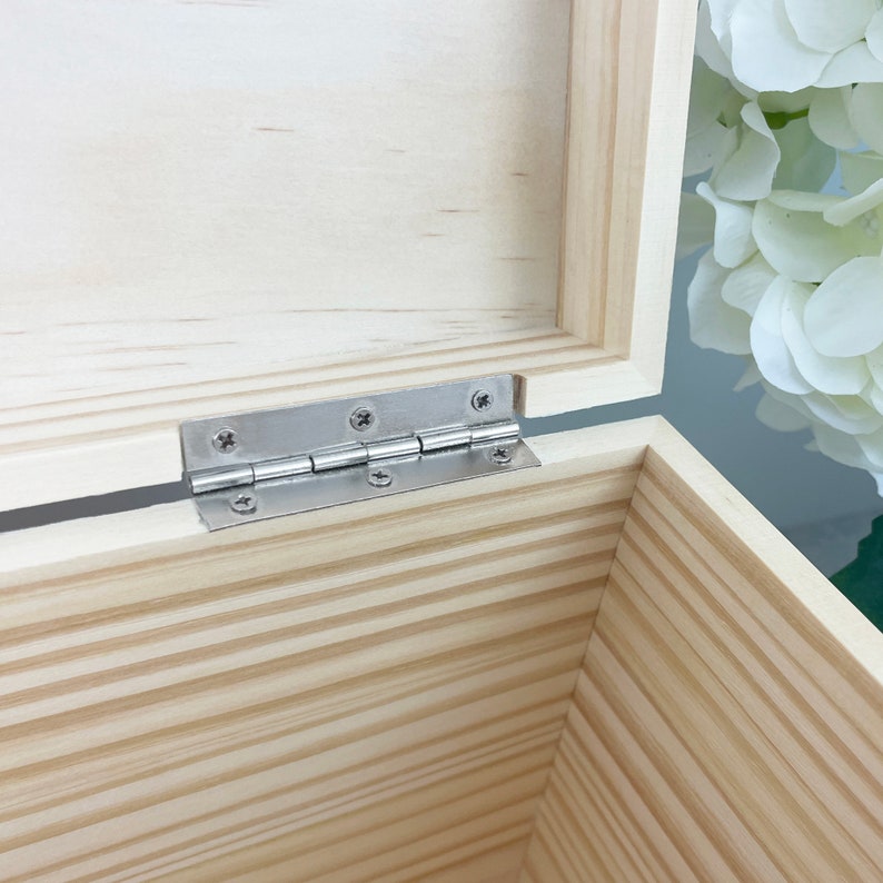 May include: An open wooden box with a light beige natural grain finish. The interior features a silver-colored metal hinge attached with four small screws, connecting the lid to the base. The box has clean, straight edges and a smooth surface.