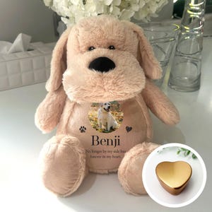 Può includere: Un giocattolo di peluche a forma di cane beige con un design a forma di zampa e cuore. Una foto di un cane è al centro del giocattolo. Il testo "Benji No longer by my side but forever in my heart." è stampato sotto la foto. Un ciondolo a forma di cuore dorato si trova nell'angolo in basso a destra dell'immagine.