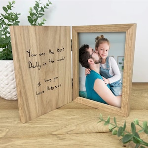 Può includere: Una cornice doppia in legno con cerniere e finitura in rovere chiaro. Il pannello sinistro è inciso con il testo: You are the best Daddy in the world, I love you, Love Ruby xxx. Il pannello destro contiene una foto di una persona e un bambino.