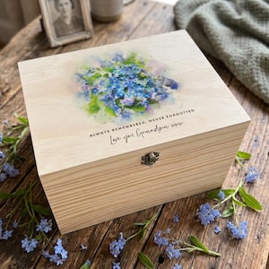 May include: A wooden box with a light-coloured finish, decorated with a watercolour floral design. The lid features blue and purple flowers and the text "Always Remembered, Never Forgotten" and "Love you Grandma xxx".