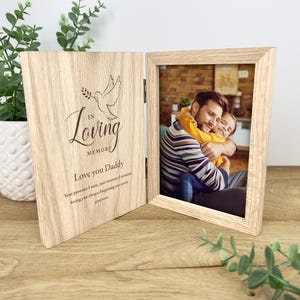 Op de afbeelding: Een houten, dubbelvouw fotolijst met een foto van een vader en kind. De linkerkant is gegraveerd met een duif en de woorden "In Loving Memory, Love you Daddy". De lijst is gemaakt van lichtgekleurd hout.