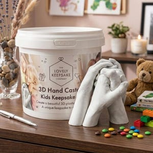 May include: A white plastic bucket with the text "THE LOVELY KEEPSAKE COMPANY" and "3D Hand Cast" is next to a white 3D hand cast. The bucket also says "Kids Keepsake" and "Create a beautiful 3D plastic". A pen, teddy bear, and colourful toys are also visible.
