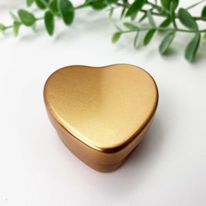 May include: A gold heart-shaped metal tin box. The box is closed and has a smooth, brushed finish.