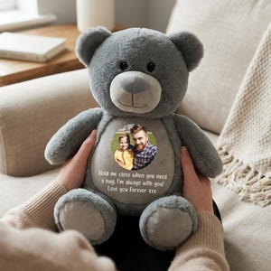 Pode incluir: Um urso de peluche cinzento com um remendo redondo no peito apresentando uma fotografia circular de uma criança e um adulto. O remendo inclui o texto impresso: Hold me close when you need a hug. I'm always with you! Love you Forever xxx.