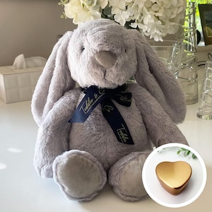 May include: A grey plush bunny rabbit with long ears, wearing a blue ribbon with gold text that reads "Teddy & Co.". The bunny is sitting on a white surface. A gold heart-shaped box is in the lower right corner of the image.