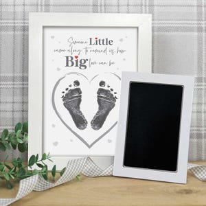 May include: A white framed print with a heart-shaped design featuring two baby footprints. The text reads, "Someone Little came along to remind us how Big love can be." A separate white frame with a black interior is also present.