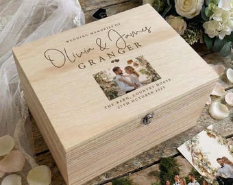 May include: A light-colored wooden keepsake box featuring the engraved text "WEDDING MEMORIES OF Olivia & James GRANGER" and a printed photograph of a couple. The box includes a metal latch and is decorated with a small heart icon above the photo.
