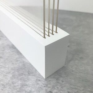 May include: A white rectangular wooden block with several vertical, thin, light-colored rods inserted into the top. The rods are evenly spaced and appear to be made of a translucent material. The block has a clean, minimalist design.