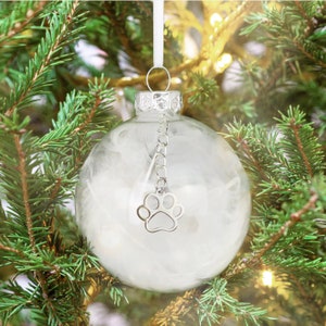 May include: A clear glass Christmas bauble with a white feather inside and a silver paw print charm hanging from a silver chain. The bauble is hanging on a green Christmas tree.