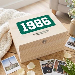 May include: A wooden memory box with the year "1986" in white text on a green rectangle. The box also has the text "40 YEARS OF MEMORIES LOVE MUM & DAD". The box is on a wooden surface with photos and flower petals.