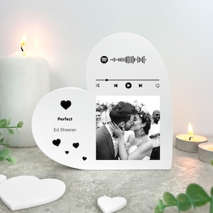 Könnte beinhalten: Ein weißer herzförmiger Fotorahmen mit einem Schwarz-Weiß-Foto eines sich küssenden Paares. Der Rahmen enthält den Songtitel "Perfect" von Ed Sheeran, Spotify-Steuerelemente und Herzgrafiken. Kerzen und Grün umgeben den Rahmen.