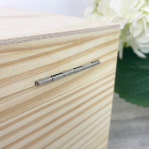 May include: A light-colored wooden box featuring a visible grain pattern and a silver-toned metal hinge connecting the lid to the main body. The box has a smooth, unfinished surface with horizontal wood grain lines running across the side panels.