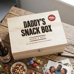 May include: A rectangular light-coloured wooden box with a metal latch. The lid features the black text DADDY'S SNACK BOX, a red DO NOT ENTER sign, and the text HAND'S OFF! BUT CAN BE SHARED WITH ELLIE & FREDDIE printed in black and red ink.