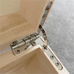 May include: Close-up of a wooden box with a partially open lid. The box is made of light-colored wood, and the hinges are shiny silver metal. The screws are also silver, contrasting with the wood.