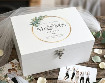 May include: A white wooden keepsake box with a gold-rimmed circular design that reads "Mr & Mrs Jones" and wedding details. The box is closed with a silver clasp. Wedding photos and floral accents surround the box.