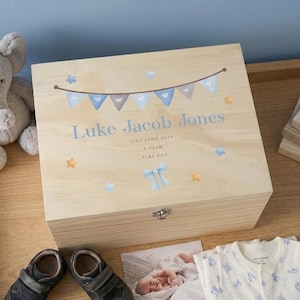 May include: A light-coloured wooden keepsake box with a hinged lid. The box is decorated with blue and grey bunting, stars, and the name "Luke Jacob Jones" in blue script. The box also includes the birth date, time, and weight: 5.32am, 3.43 kg.