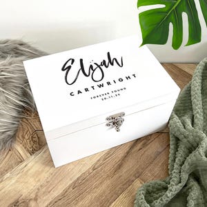 Könnte beinhalten: Eine weiße Holzkiste mit silbernem Verschluss. Der Deckel trägt den Namen "Elijah" in Schreibschrift, zusammen mit dem Namen "Cartwright" und dem Satz "Forever Young 20.11.25" in einer einfachen Schriftart.