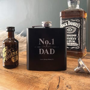 Può includere: Una fiaschetta nera con la scritta "No. 1 DAD" in lettere argentate. Una bottiglia di whisky Jack Daniel's e una piccola bottiglia di rum speziato sono sullo sfondo. È presente anche un imbuto d'argento.
