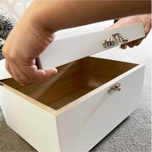 May include: A white wooden box with a hinged lid is being opened. The box has a silver clasp and hinges. The interior of the box is natural wood. The box is rectangular and appears to be empty.