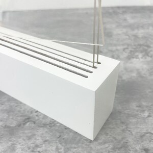 May include: A white wooden knife block with several slots for knives. The block has a rectangular shape and is positioned at an angle. A clear acrylic or glass panel is visible above the block, with thin metal rods extending from it.