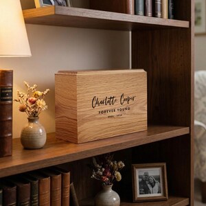 Könnte beinhalten: Eine hellbraune Holzurne mit der Aufschrift "Charlotte Cooper Forever Young 2006-2020" steht auf einem Holzregal. Das Regal enthält auch Bücher, eine kleine Vase mit Blumen und ein gerahmtes Foto. Die Urne ist rechteckig.