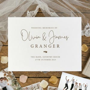 May include: A white wedding memory box with the text "WEDDING MEMORIES OF Olivia & James GRANGER" in brown script. The box also includes the date "27TH OCTOBER 2023". The box is surrounded by wedding photos and flower petals.
