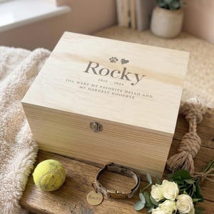 Puede incluir: Una caja conmemorativa de madera clara para mascotas con el nombre "Rocky" y las fechas 2015-2024 grabadas en la tapa. Una pelota de tenis, un collar para perros y flores están junto a la caja. El texto dice: "Fuiste mi hola favorito y mi adiós más difícil."