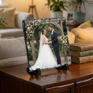 Könnte beinhalten: Rechteckiger Foto-Stein-Druck mit einem Hochzeitsfoto eines Paares, das sich unter einem Blumenbogen an den Händen hält. Der Druck wird auf einem schwarzen Ständer auf einer Holzoberfläche präsentiert.