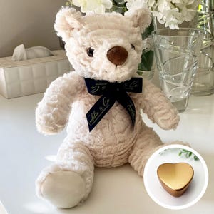 May include: A white plush teddy bear with a brown nose and a blue ribbon with the text "Cuddles & Co" in gold lettering. The bear is sitting on a white surface with a gold heart-shaped box in the background.