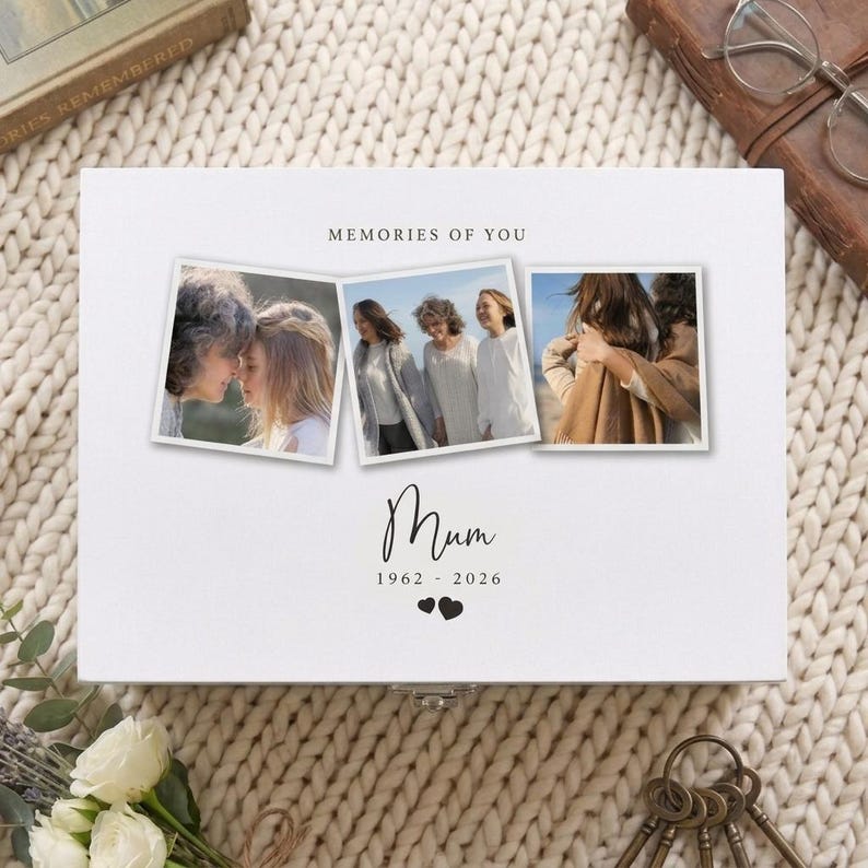 May include: A white keepsake box with three framed photos. The box is labeled "MEMORIES OF YOU" and features the word "Mum" with the years 1962-2026. Two small black hearts are below the dates. A metal clasp is visible.