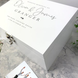 May include: A white wooden memory box with silver hardware. The box is inscribed with the words "Wedding Memories of Olivia & James Granger, The Barn, Country House, 27th October 2023" in gray script. A photograph is visible in the foreground.