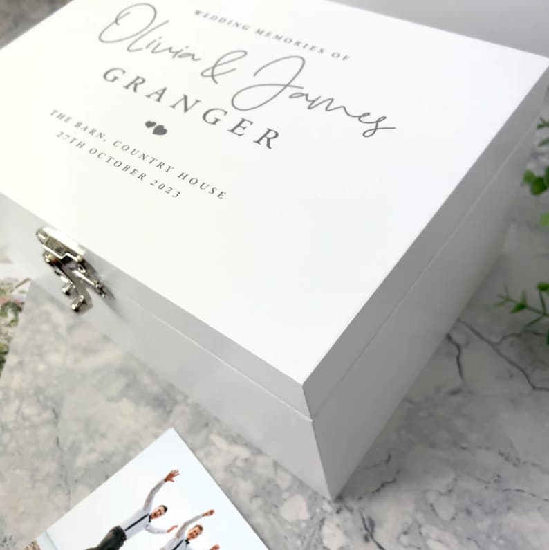 May include: A white wooden memory box with silver hardware. The box is inscribed with the words "Wedding Memories of Olivia & James Granger, The Barn, Country House, 27th October 2023" in gray script. A photograph is visible in the foreground.