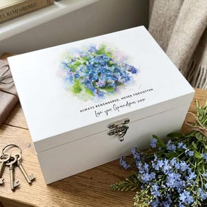 May include: A white wooden memory box with a floral watercolour design on the lid. The design features blue and purple flowers with green accents. Text on the lid reads "ALWAYS REMEMBERED, NEVER FORGOTTEN" and "Love you Grandma xxx". The box has a metal clasp.